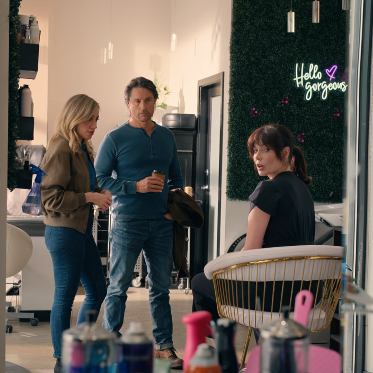 Two people standing in a salon with a third person sitting in a chair, neon sign in the background.